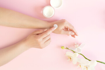 Well-groomed female hands with cosmetic cream on a pink background. A sprig of orchid and a jar of cream.