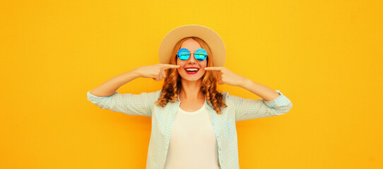 Portrait of happy smiling caucasian young woman pointing her fingers at her teeth wearing straw hat on orange background