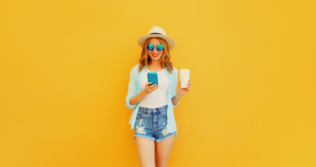 Happy smiling young woman 20s with smartphone and cup of coffee wearing summer straw hat, shorts on orange background