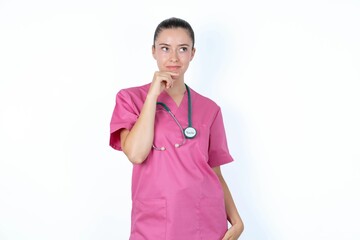 Shot of contemplative thoughtful young caucasian doctor woman wearing pink uniform over white background keeps hand under chin, looks thoughtfully upwards.