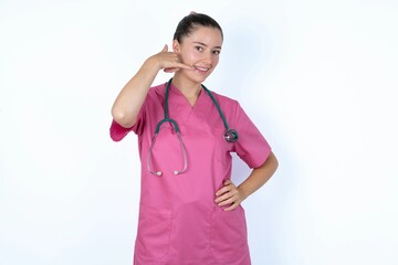 young caucasian doctor woman wearing pink uniform over white background imitates telephone conversation, makes phone call gesture with hands, has confident expression. Call me!