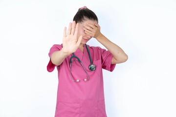 young caucasian doctor woman wearing pink uniform over white background covers eyes with palm and doing stop gesture, tries to hide. Don't look at me, I don't want to see, feels ashamed or scared.