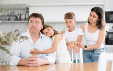 Saddened by bad news, man is sitting in kitchen surrounded by sympathetic family. Child hugs distressed dad, wife and daughter stand behind man