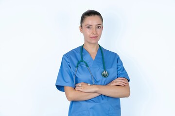 Self confident serious calm young caucasian doctor woman wearing medical uniform over white background stands with arms folded. Shows professional vibe stands in assertive pose.