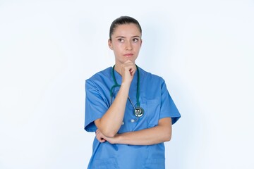 Thoughtful young caucasian doctor woman wearing medical uniform over white background holds chin and looks away pensively makes up great plan