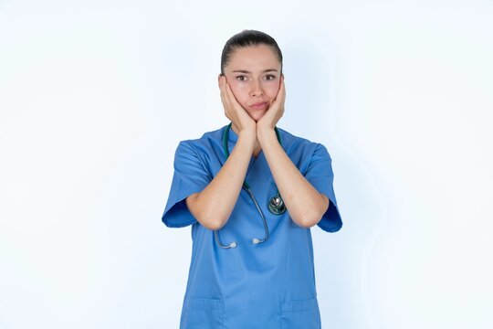 young caucasian doctor woman wearing medical uniform over white background with surprised expression keeps hands under chin keeps lips folded makes funny grimace