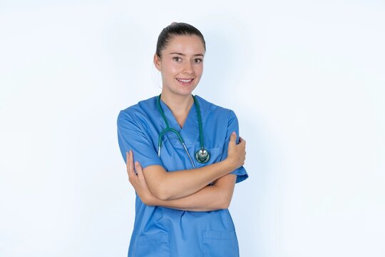 Charming pleased young caucasian doctor woman wearing medical uniform over white background embraces own body, pleasantly feels comfortable poses. Tenderness and self esteem concept