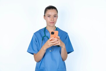 Portrait of serious confident young caucasian doctor woman wearing medical uniform over white background holding phone in two hands