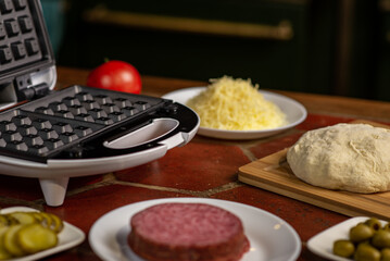 Cooking pizza waffle, piffle. Different ingredients - sausage, olives, pickles, dough on the kitchen table.