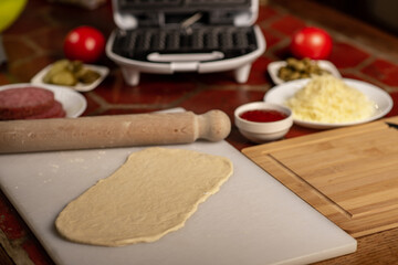 Cooking pizza waffle, piffle. Waffle iron and yeast dough for pizza and various ingredients - salami, cheese, olives, tomato on the kitchen table.