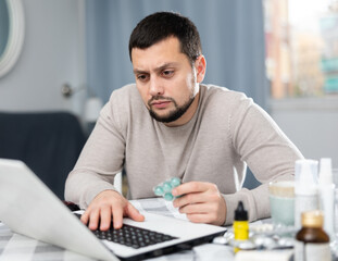 Interested young man looking for information about medicines on Internet using laptop while sitting at table at home..