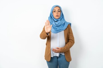 Young beautiful pregnant muslim woman wearing hijab over white background shows stop sign prohibition symbol keeps palm forward to camera with strict expression