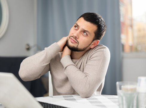 Tired young man massaging neck muscles while suffering from pain after long work on computer at home..