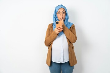 Young beautiful pregnant muslim woman wearing hijab over white background holding modern gadget ask...