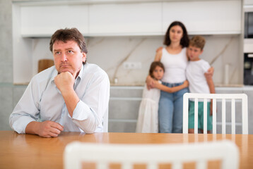 Fototapeta premium Thoughtful father is sitting in kitchen and thinking about ways to solve difficult financial situation. Wife and children stand behind dad and are afraid to ask about current problem