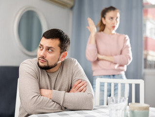 Obraz premium Sad man sitting at table in apartment while his wife standing behind and quarreling with him.