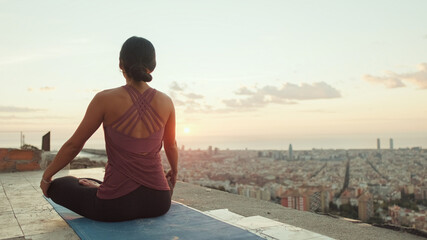 Fototapeta na wymiar Young woman in bodysuit practices yoga at sunrise at viewpoint. Back view