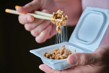 Hand holding chopsticks with natto fermented soy beans in styrofoam box.