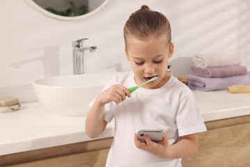 Little girl using smartphone while brushing teeth in bathroom, space for text. Internet addiction