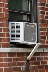 vertical shot of exterior view of air conditioning window unit extruding from the window sill of a red brick building with a portable safety ledge support installed under it and