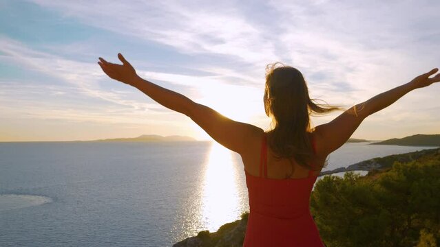 LENS FLARE, CLOSE UP: Summer breeze ruffles woman hair as she raises her arms. She is fascinated by the beautiful morning sun rising over blue Adriatic Sea. Young lady enjoying on holidays at seaside.