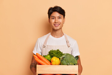 Smiling guy in white T-shirt posing against studio background holding wooden box full of vegetables, organic food concept, copy space