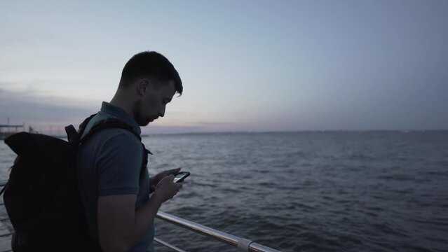 Young caucasian man taking photo on smartphone while relaxing on pier near sea evening. Male taking cell phone picture of sunset over the black sea. Evening scenery near sea using mobile phone camera.