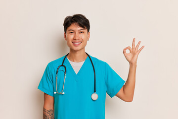 Young male doctor in medical uniform poses on white background, smiling and showing with his fingers OK sign, business concept, copy space
