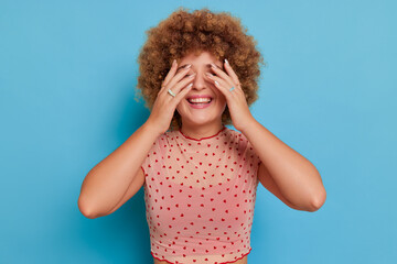 Happy girl with afro hairstyle posing on blue background, stands smiling and closing eyes with hands, fun time concept, copy space