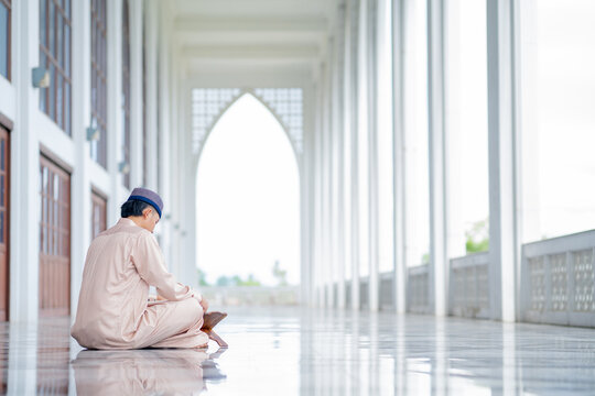  An Asian Muslim man is sitting and reading the Quran. The peace in the mosque makes it an energetic atmosphere of faith, with copy space,  Arabic word translation: The Holy Al Quran.