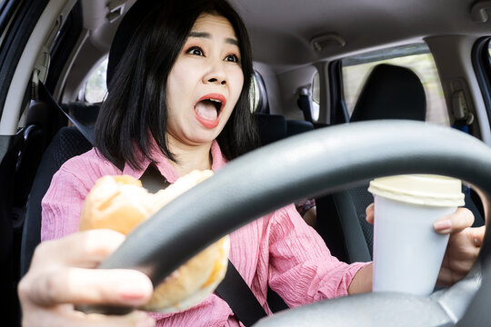 dangers of distracted eating while driving concept with careless Asian woman's car accident with food and drink in hand on steering wheel