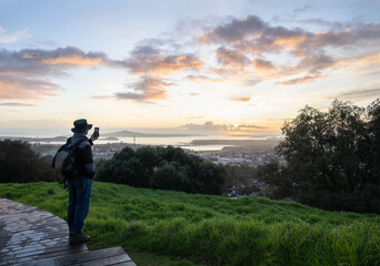 Tourist taking photo using smartphone at Mt Eden summit at sunrise. Rangitoto Island in the distance. Auckland.