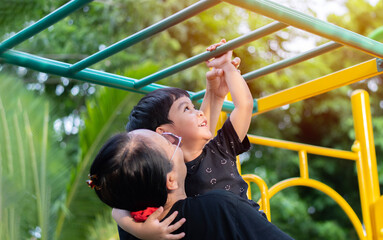 Happy Asian boy try to hang the green bar by his mother holding and helping him at out door playground