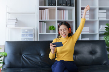 Happy young Asian woman raising hands with victory smiling happily with smart phone at home. Success, win, victory, triumph, congratulation, concept..