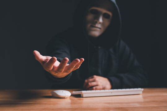 Black hood hacker force demands ransom by holding out his hand on table where sensitive data is hacked in a dark room in the background. Cyber security and cyber crime concept. Hacking and phishing