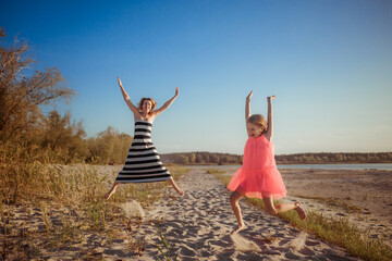 Happy mother and daughter in dresses are jumping and dancing on the beach during sunset. A good relation between generations. Fun family pastime. Health promotion through outdoor activities. Vacation