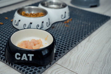Pieces of chicken raw meat is on kitten bowl with cat inscription standing on mat on wooden floor...