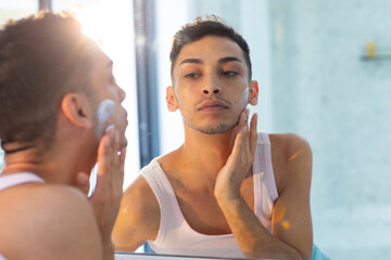 Biracial transgender man looking in mirror and applying face cream in bathroom