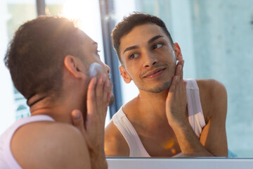 Happy biracial transgender man looking in mirror and applying face cream in bathroom