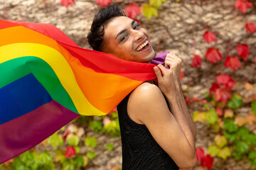 Portrait of happy biracial transgender man holding rainbow flag, with rocks and leaves in background