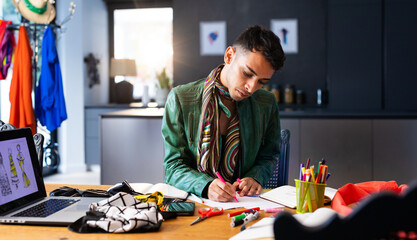 Biracial transgender fashion designer sitting at desk, using laptop and drawing sketches