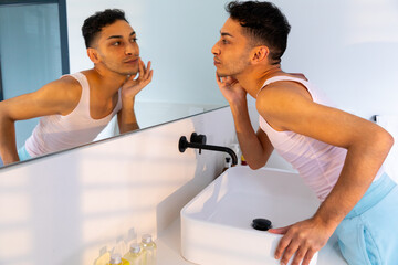 Biracial transgender man looking in mirror and touching face in bathroom