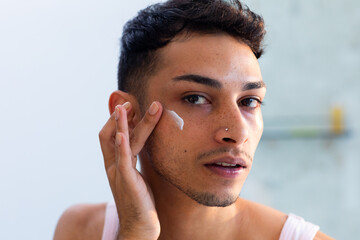 Biracial transgender man looking in mirror and applying face cream in bathroom