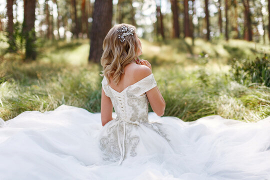 beautiful bride enjoy on nature back view. blonde woman in long white dress sitting on green grass in summer park outdoor portrait. wedding day