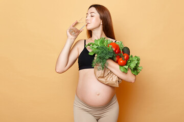 Water balance and healthy nutrition during pregnancy. Healthy pregnant woman embraces fresh vegetables posing isolated over beige background holding glass drinking water.