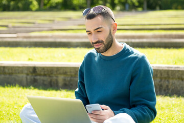 Fototapeta premium Young bearded man sitting in a garden with laptop and phone