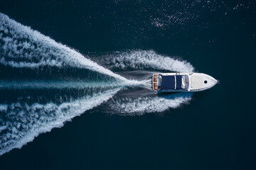 A white large boat with a blue awning is moving fast on dark water, top view.