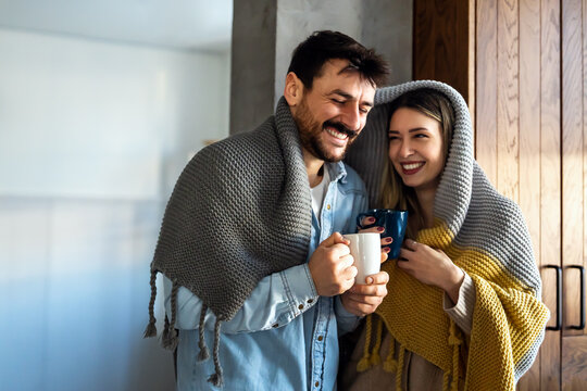 Happy Young Couple With Hot Drinks Cuddling Under Warm Blanket At Home