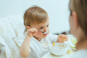 Little toddler eating using spoon in hand, learning how to feed himself. Infant sitting on child chair, eating from white bowl, trying healthy food lunch time at home baby led weaning