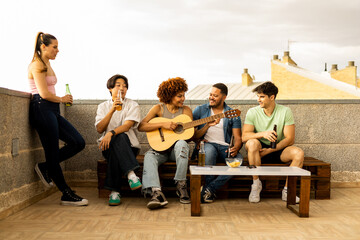 A group of multi-ethnic youths are having fun on a rooftop of an outdoor floor. The girl with afro hair plays a classical guitar while the others drink bottles of beer.Concept of Spanish culture.